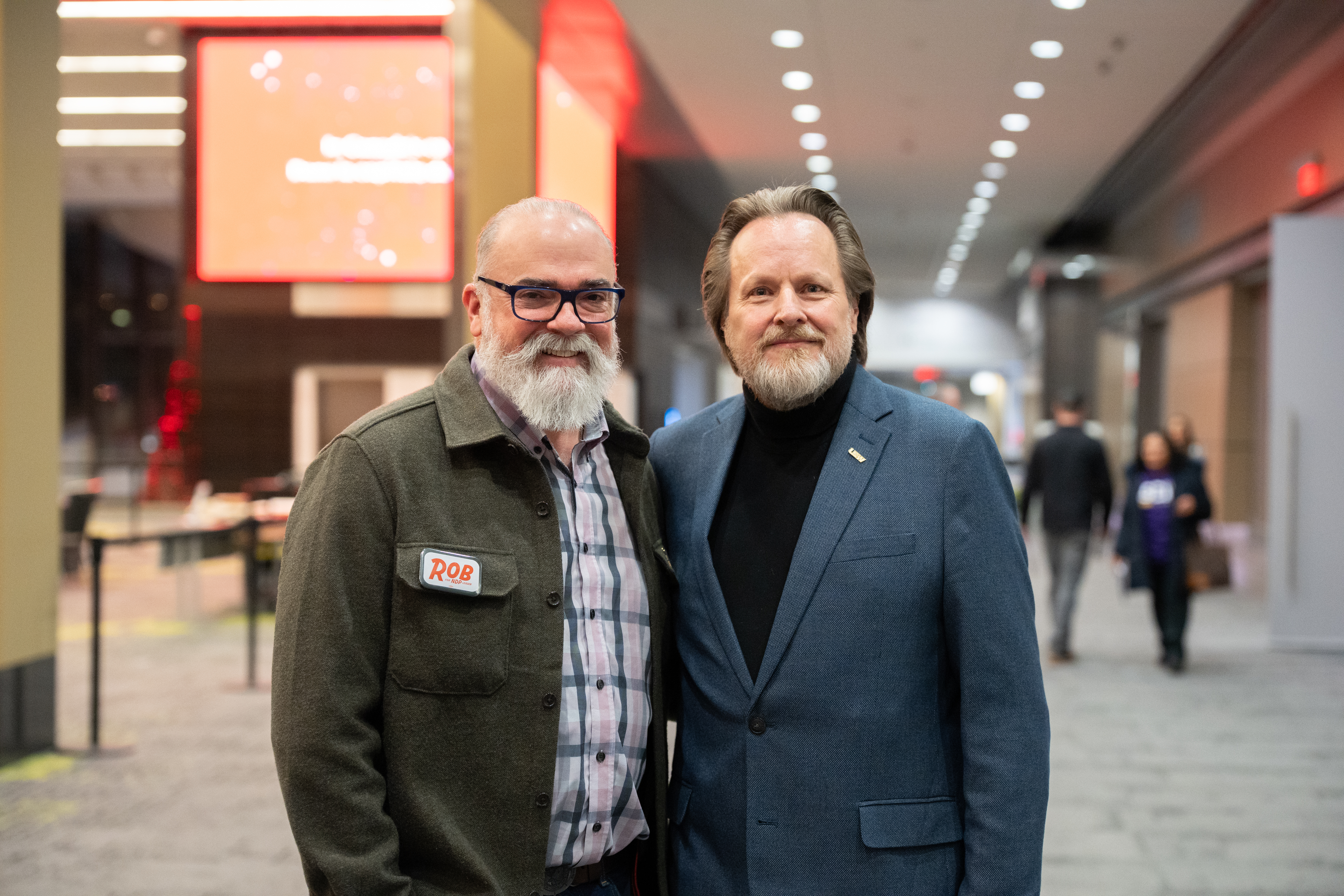 Two men are shown from the waist up standing side by side in the halls of a convention centre. Both are bearded and the one on the left is wearing gla