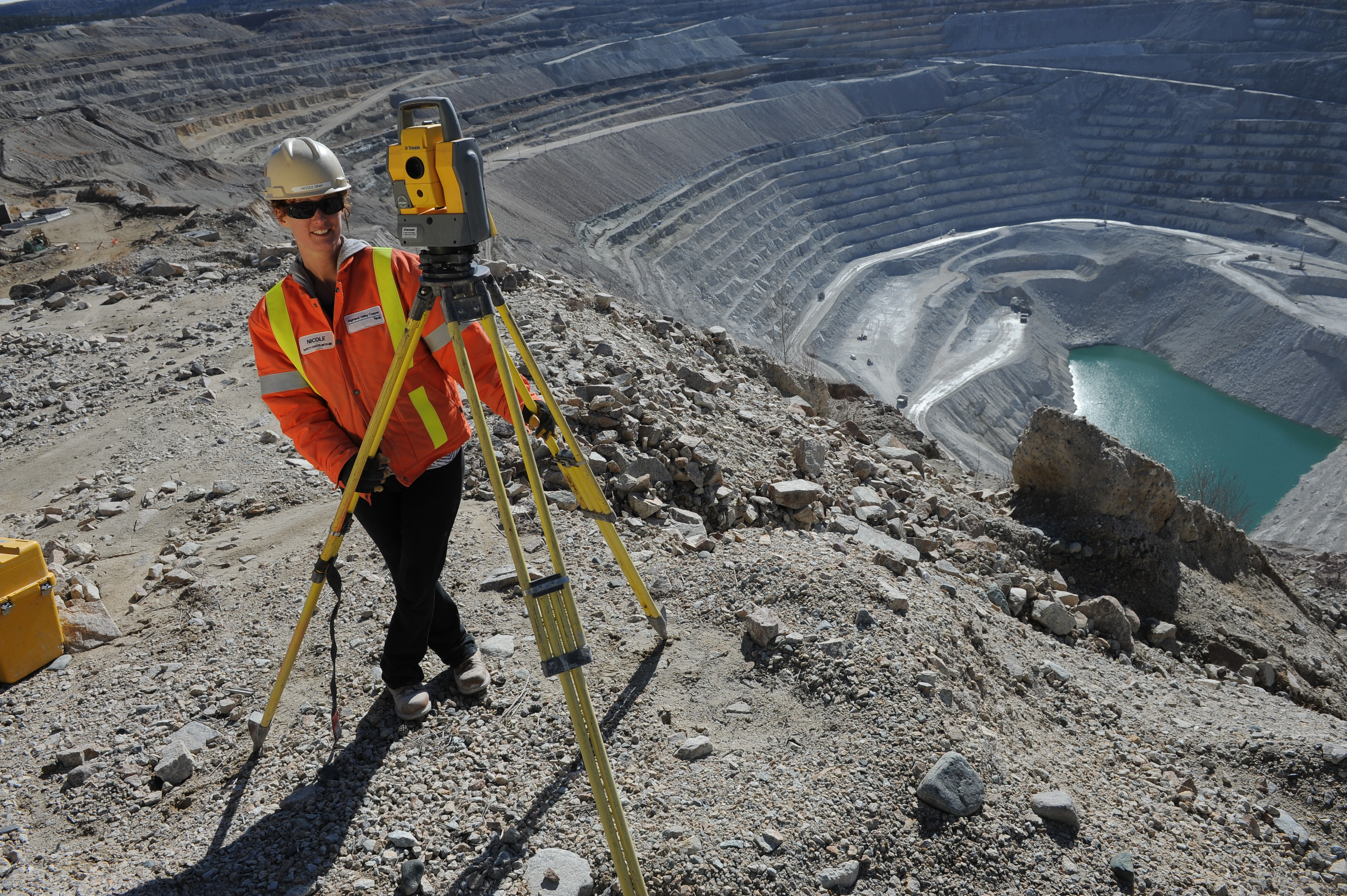 A person in a reflective vest is using survey equipment. Behind them is a deep valley with a surface copper mine and water at the bottom is visible.